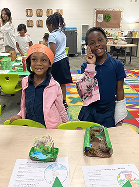 Two little African-American girls smiling at the camera and standing by tables with projects on them